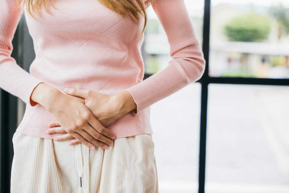 woman in pink sweater and sweatpants touching her lower abdomen because of bloating, stomach pain